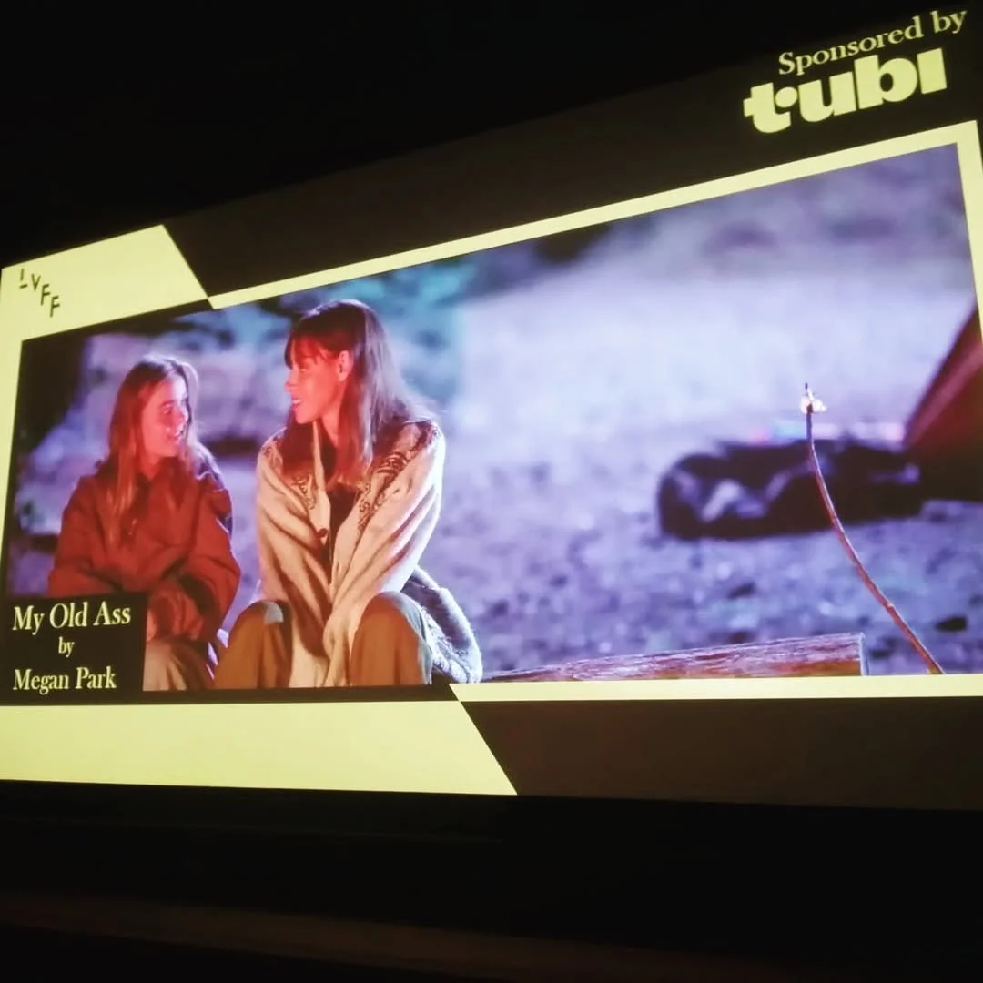 Scene from a movie or show featuring two women sitting outdoors in a nighttime setting. The woman on the left has long hair and is wearing a brown jacket, while the woman on the right has shorter hair and is wearing a patterned jacket. There is text 