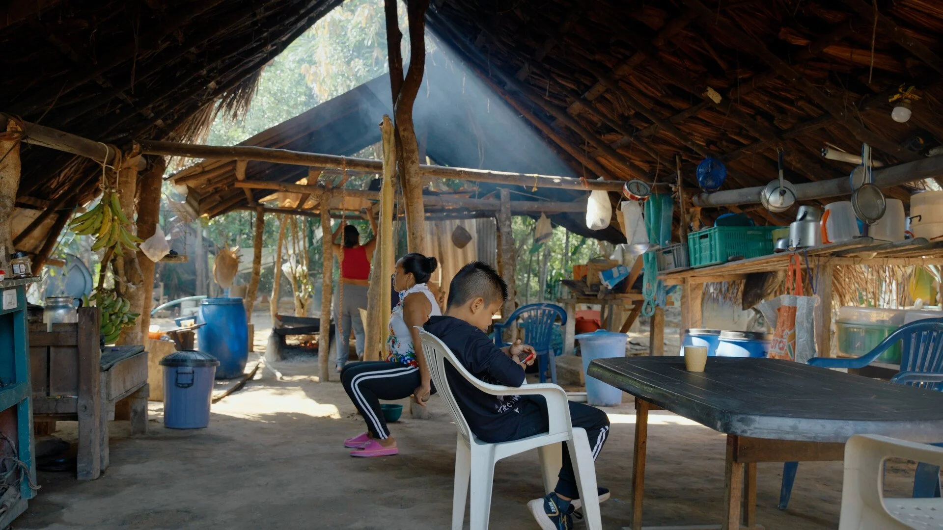 Children playing inside a rustic open-air kitchen or shelter with a thatched roof and hanging utensils, with sunlight filtering through trees.