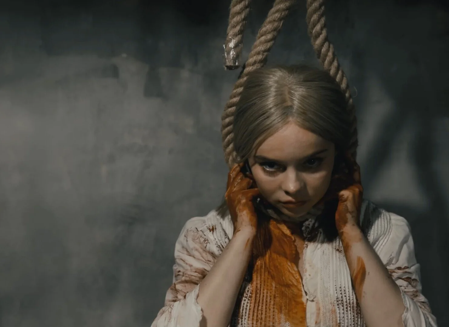 A young girl with blond hair, hanging from a noose with her hands on her ears, covered in blood, against a dark gray wall.