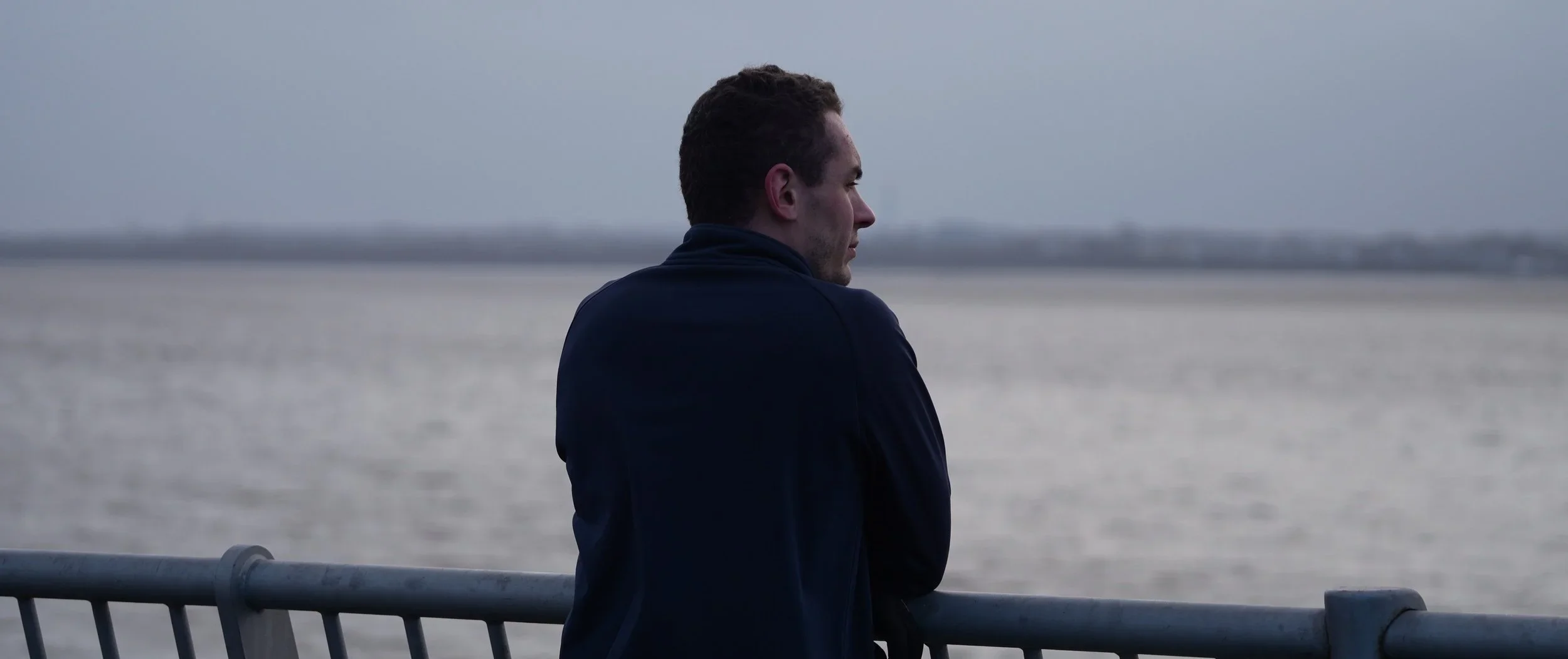 A man with short, curly hair stands near a metal railing, gazing out at a body of water on a cloudy day.