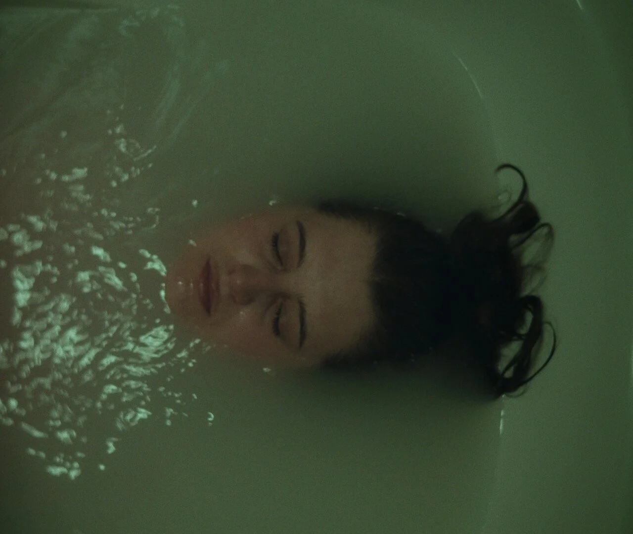 A woman with dark curly hair floating in water with her eyes closed, surrounded by bubbles, and appearing relaxed.