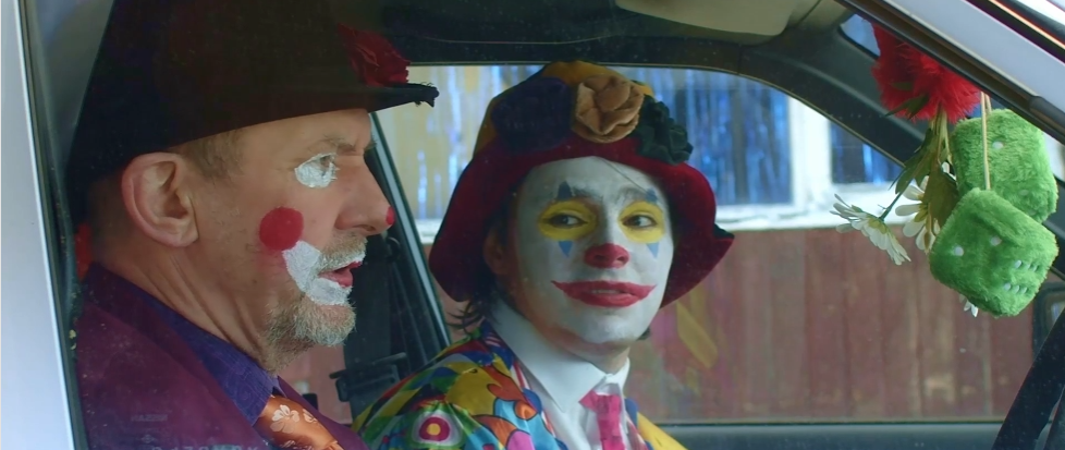 Two clowns, one older and one younger, sitting inside a vehicle, with clown makeup and colorful costumes, surrounded by clown-themed decorations hanging from the mirror.
