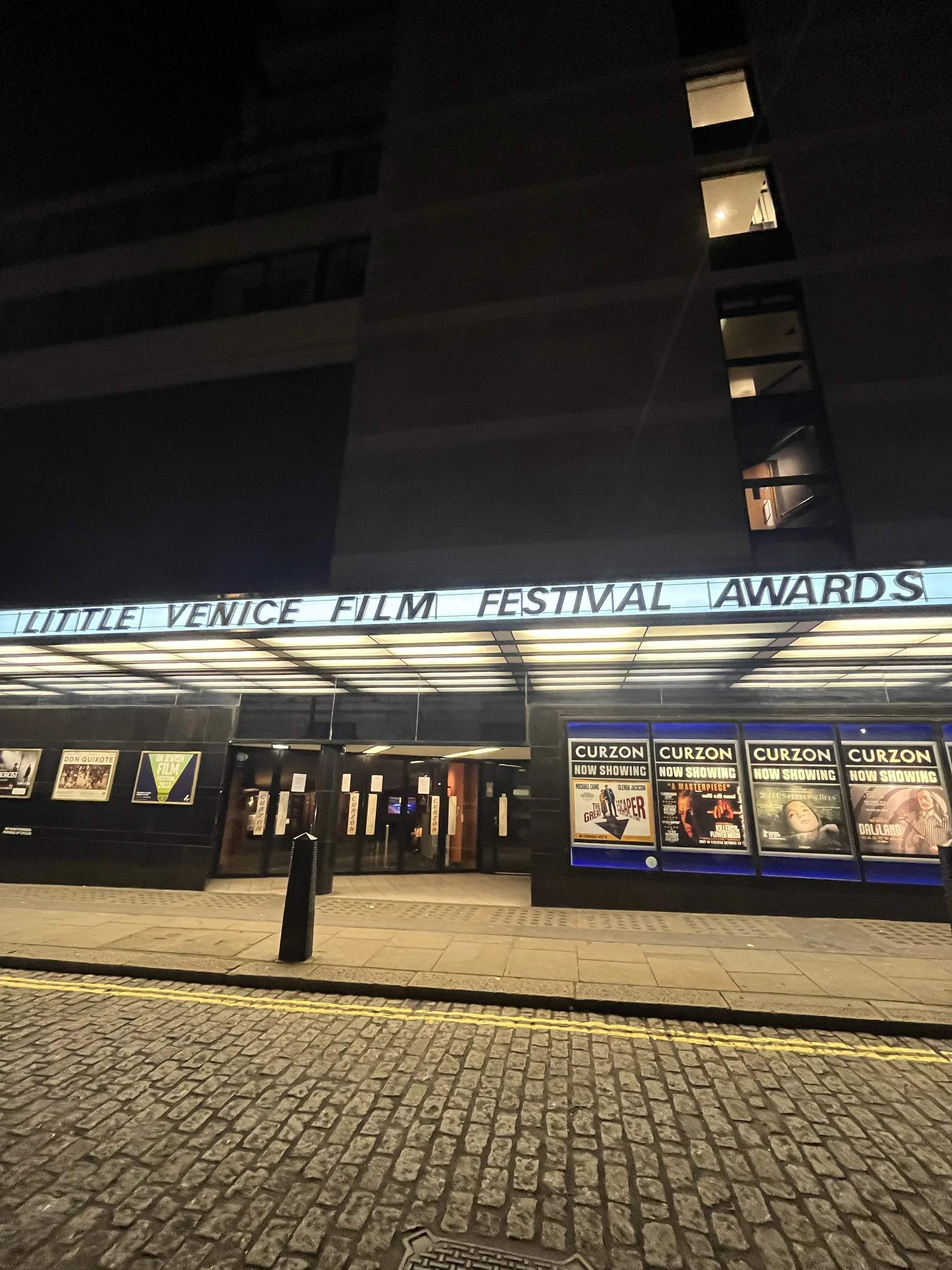 The entrance of the Little Venice Film Festival Awards in a building at night, with a marquee sign displaying the event's name and posters advertising movies being shown.