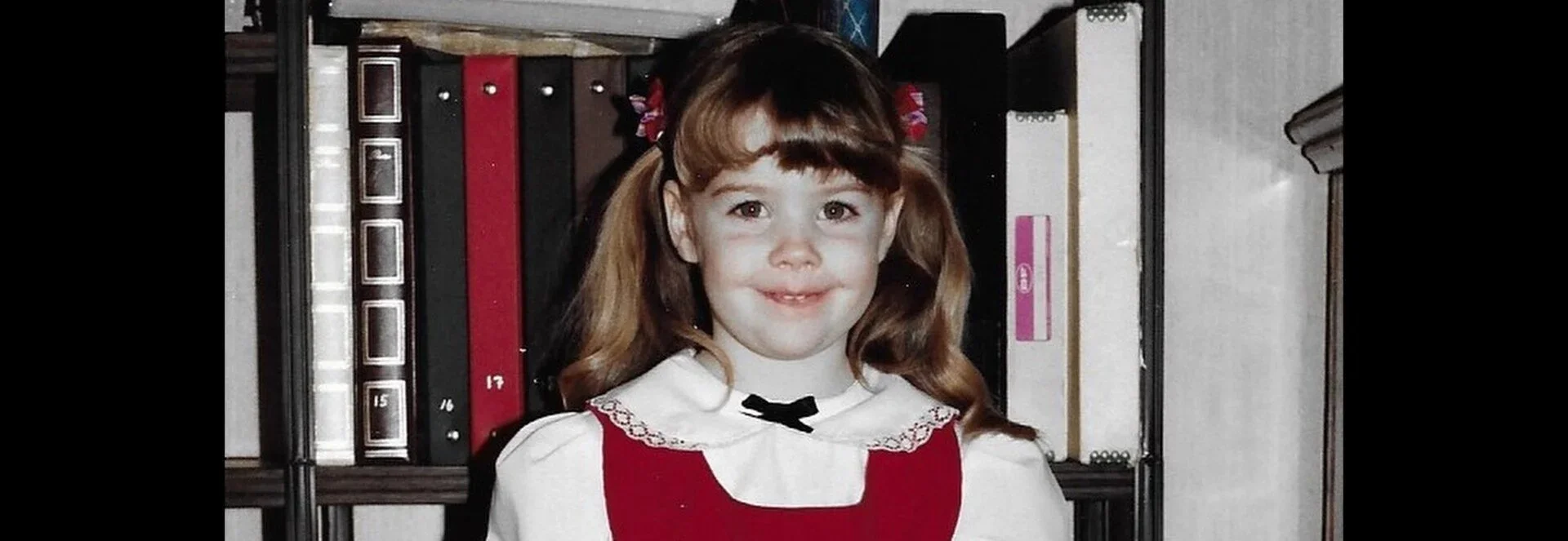 A young girl with brown hair styled in pigtails, wearing a white blouse with a black bow and a red dress, standing in front of a bookshelf.