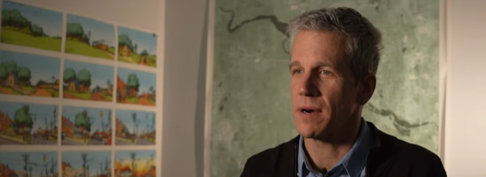 A man with gray hair stands indoors in front of a wall decorated with colorful landscape artworks and a large map or painting.