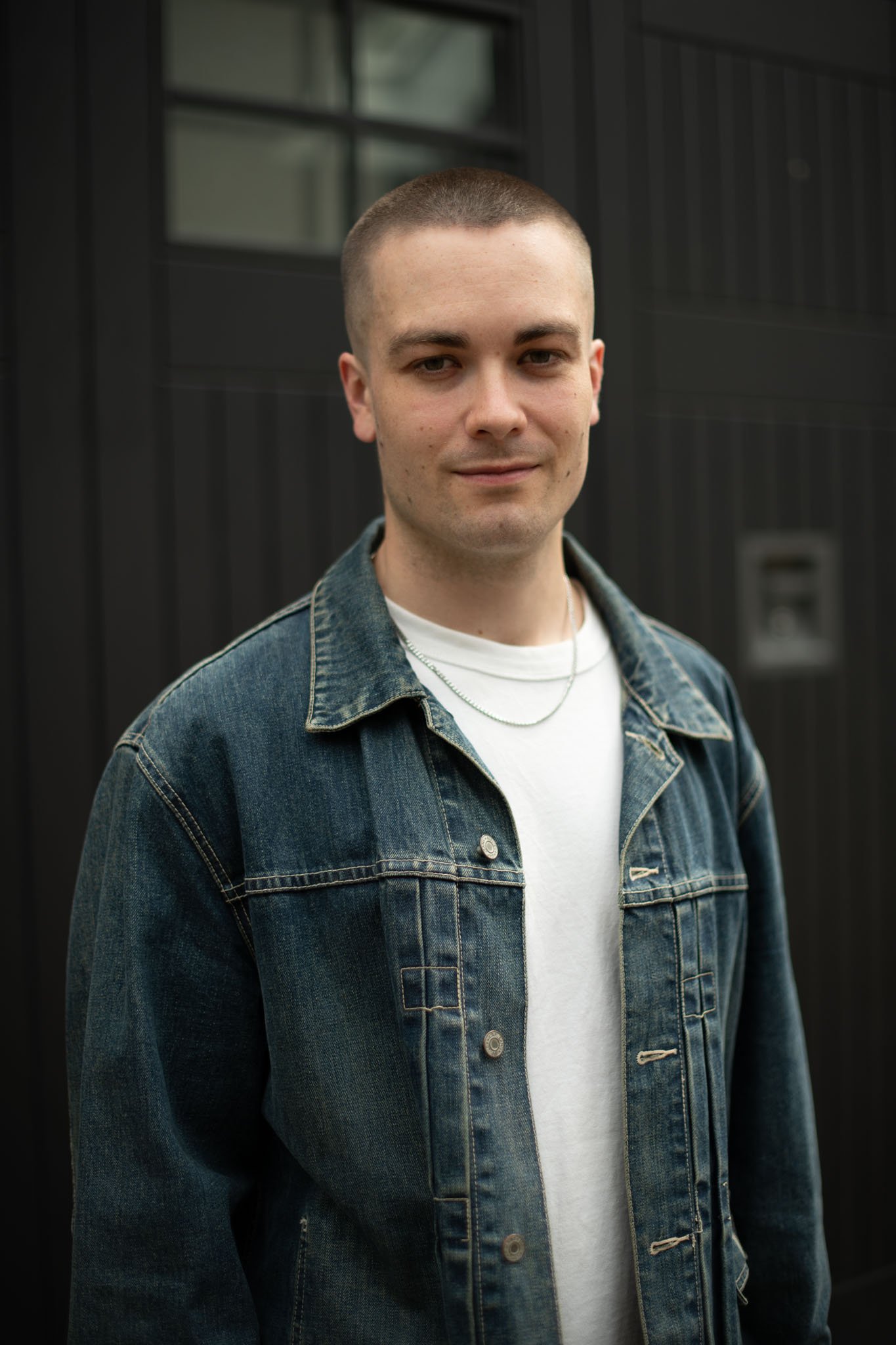 A young man standing outdoors in front of a black building wearing a denim jacket and a white T-shirt.