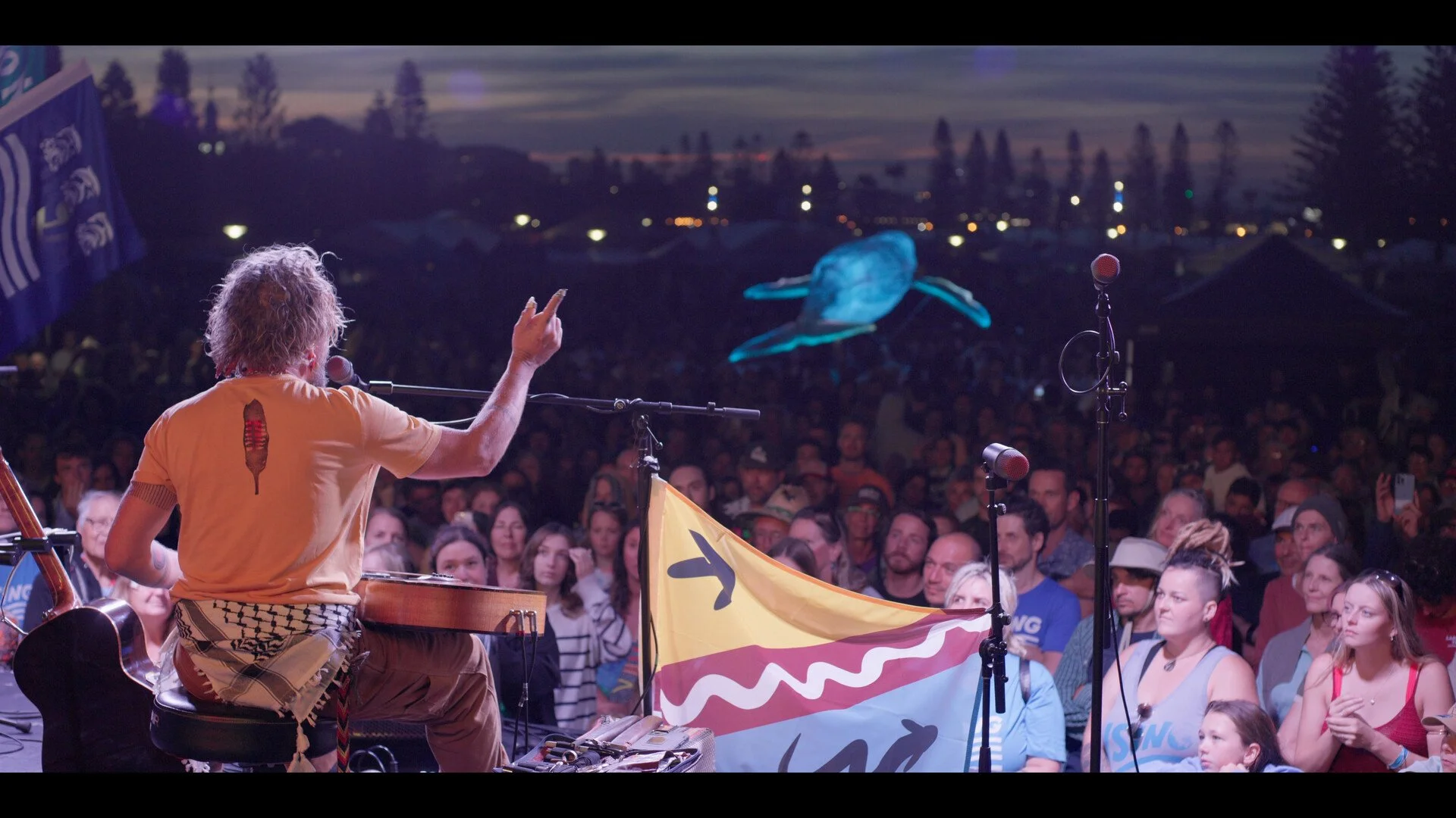 A musician with curly hair, wearing a yellow T-shirt with a chili pepper design, performs on stage with a guitar and a microphone in front of a large audience at dusk. The crowd listens attentively, and a glowing blue whale sculpture is visible in the background.