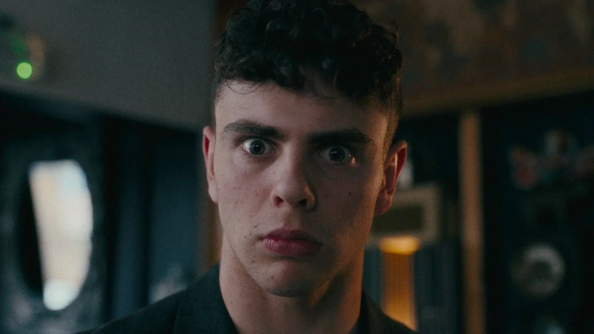 A young man with curly dark hair and pale skin stares directly at the camera with wide eyes and a serious expression. The background appears to be a dimly lit room with speakers and electronic equipment.