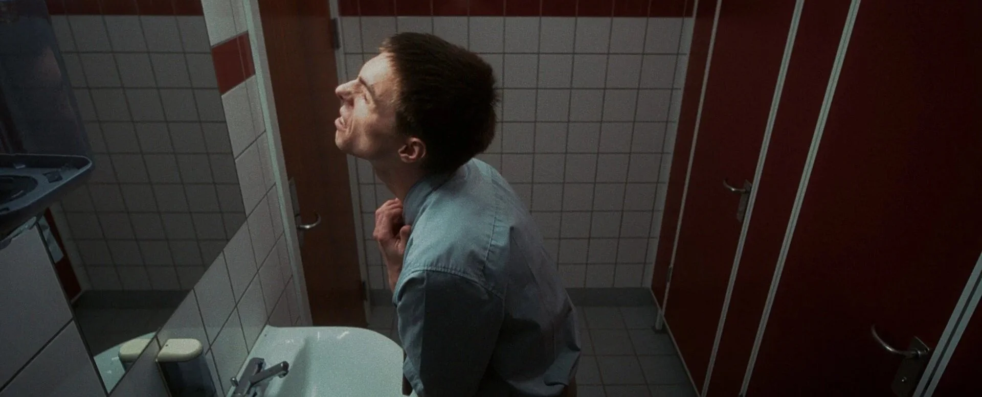 A man in a public restroom wearing a light shirt, looking upwards, with a tense expression, standing near a sink, with bathroom stalls in the background.