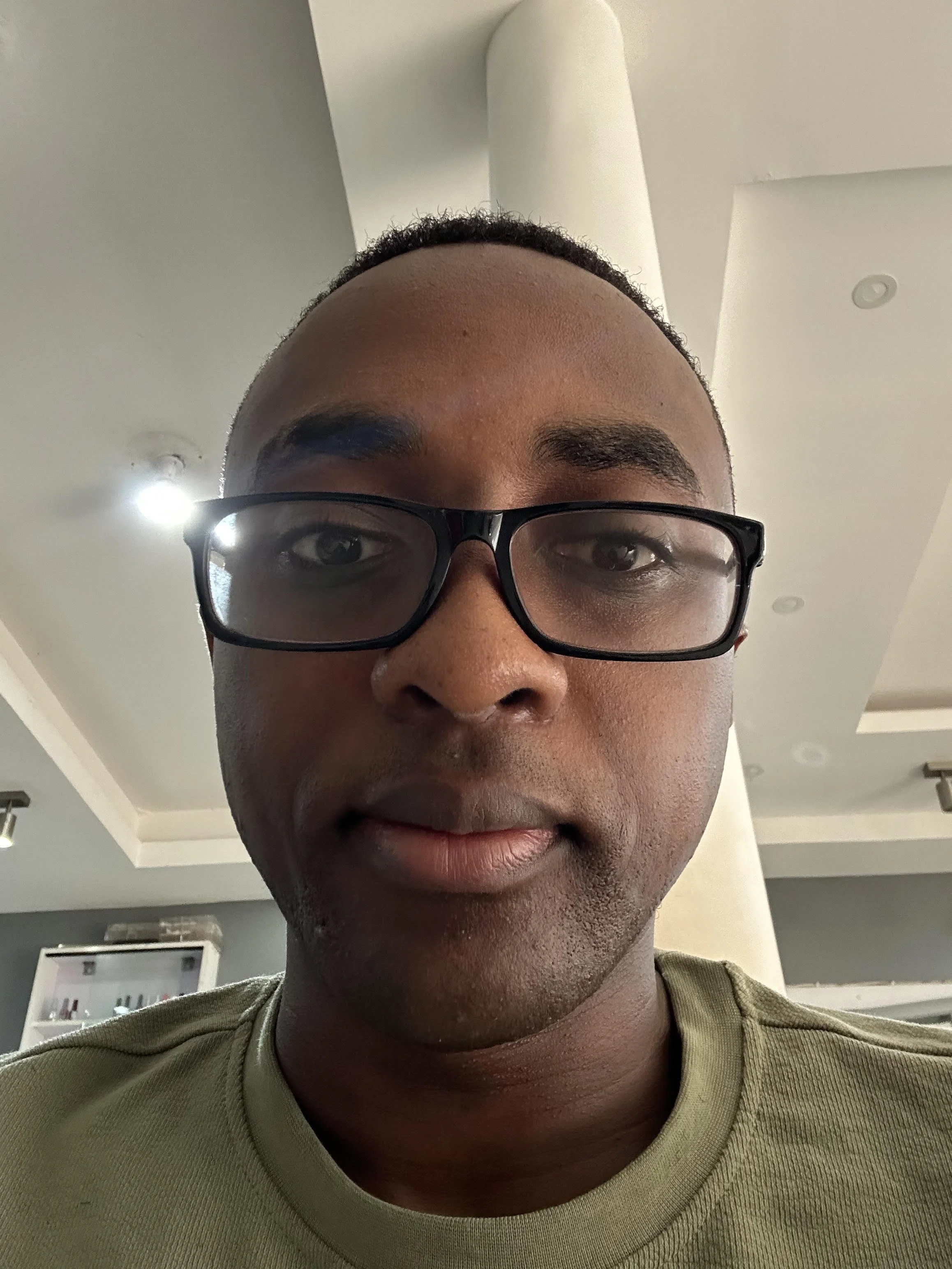 Close-up of a man wearing black glasses and a khaki shirt, indoors with a white ceiling and light fixtures in background.