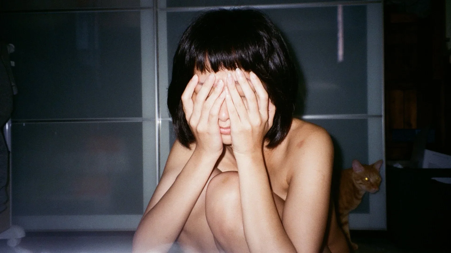 A topless woman with short black hair covering her face with her hands, sitting on the floor, with a cat peeking out from behind her. The background includes a gray cabinet and a dark room.