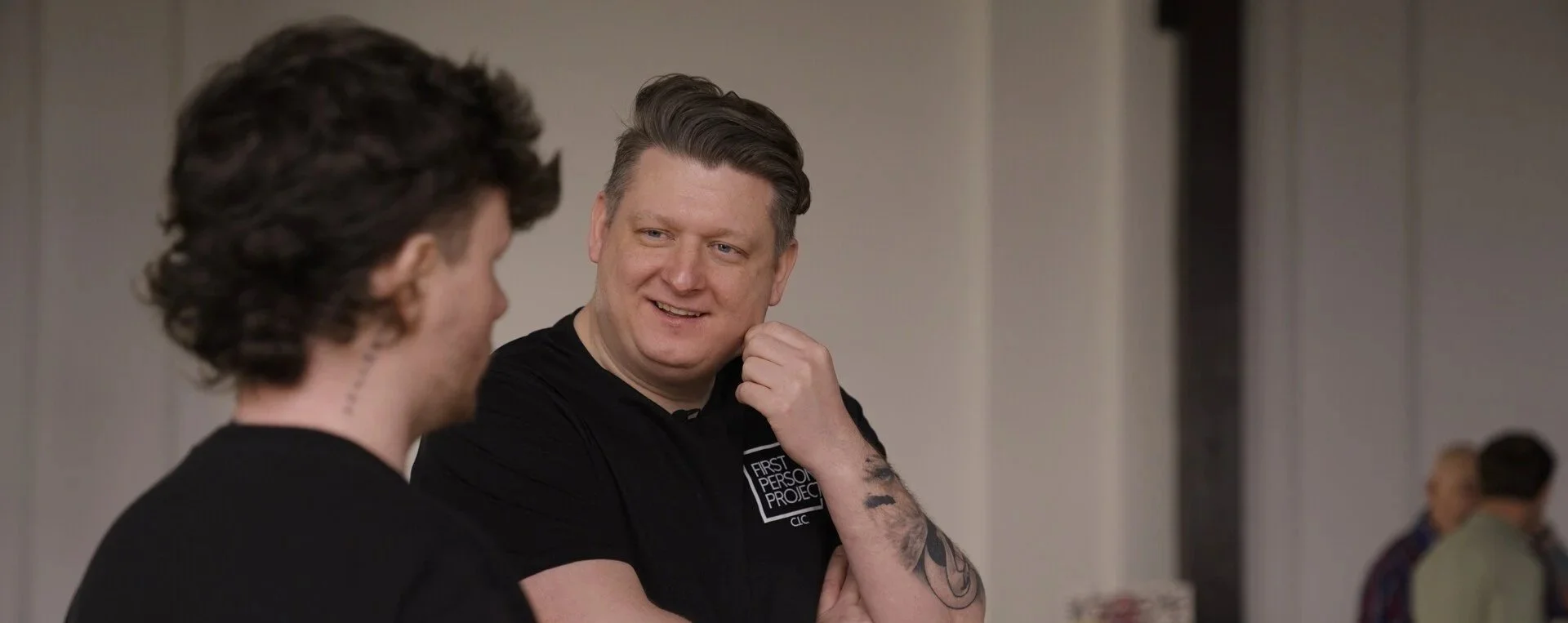 Two men engaged in conversation, smiling, in an indoor setting, with others in the background.