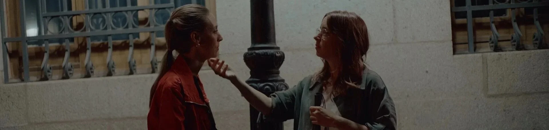 Two women are standing outside at night, engaged in a serious conversation. The woman on the left has blonde hair tied back, and the woman on the right has red hair and glasses. They are near a stone wall with a metal fence and a lamppost.