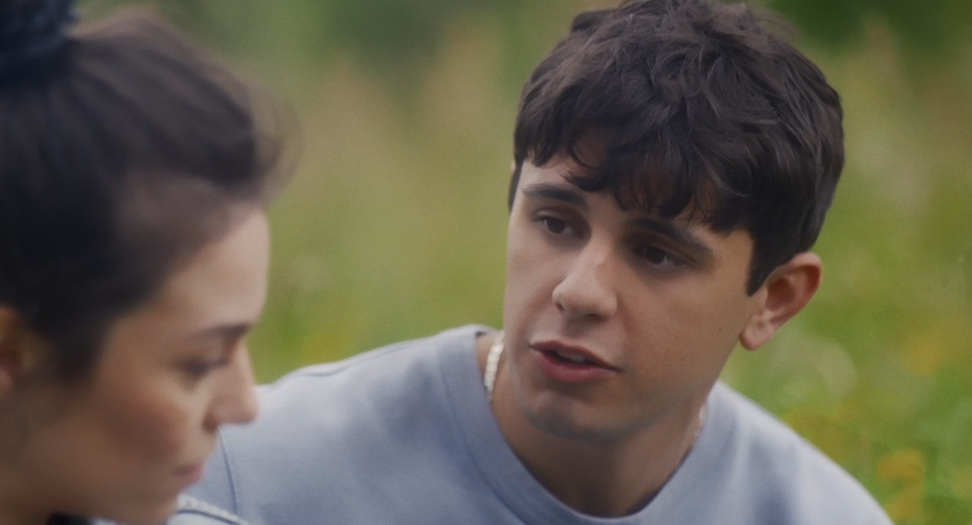 A young man talking to a woman outdoors with greenery in the background.
