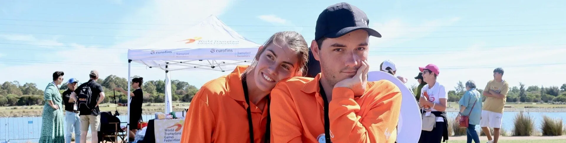 Two young adults, one smiling and one looking pensive, wearing orange shirts and black lanyards, at an outdoor event with a tent, a lake, and other people in the background.