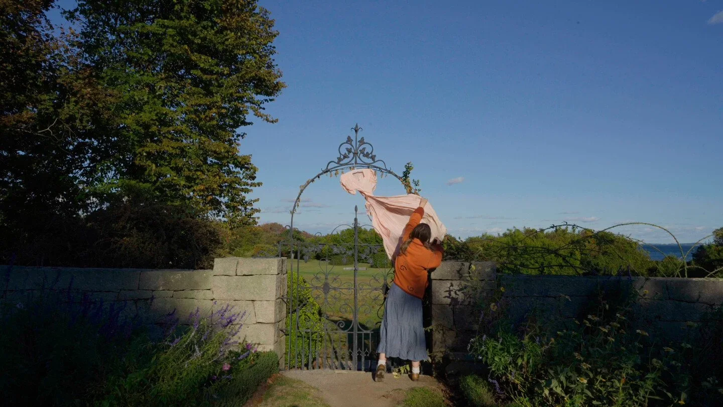 A person wearing an orange top, denim skirt, and a white cap standing at an iron gate, holding back a curtain or cloth, with a stone wall and green trees in the background.