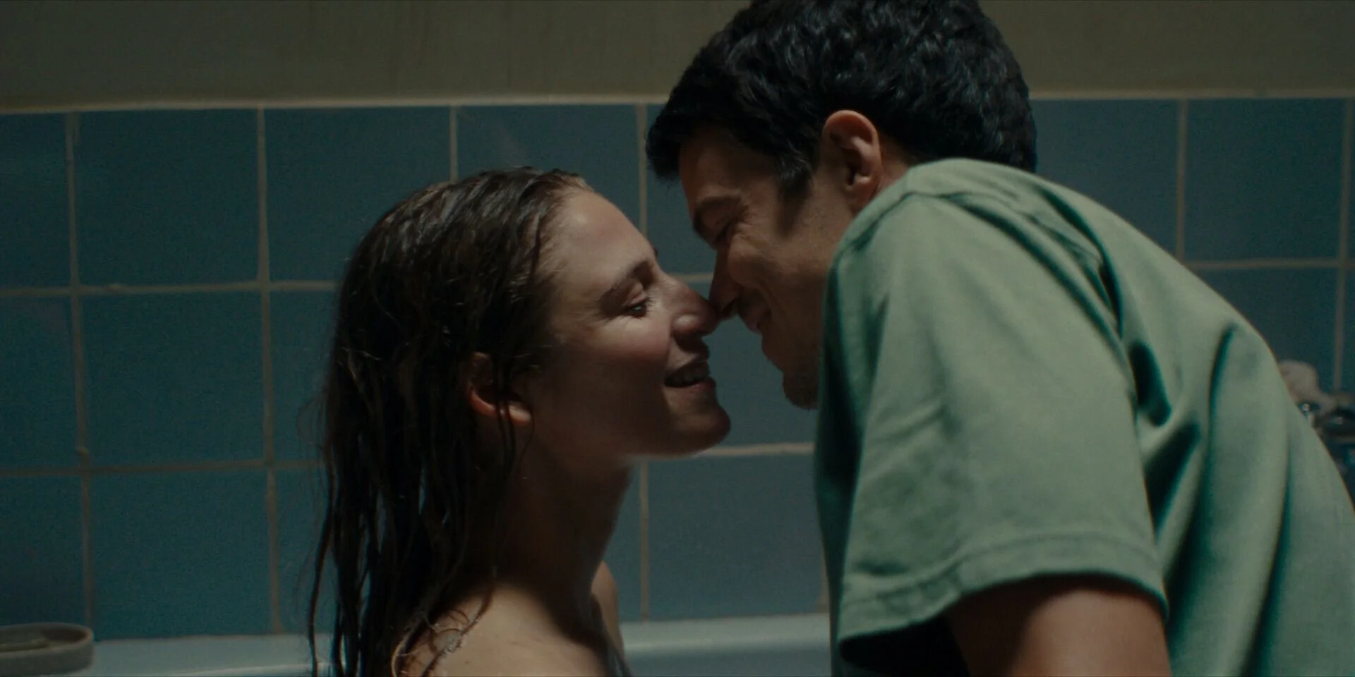 A man and woman in a close, intimate moment, touching faces in a bathroom with blue tiled walls.