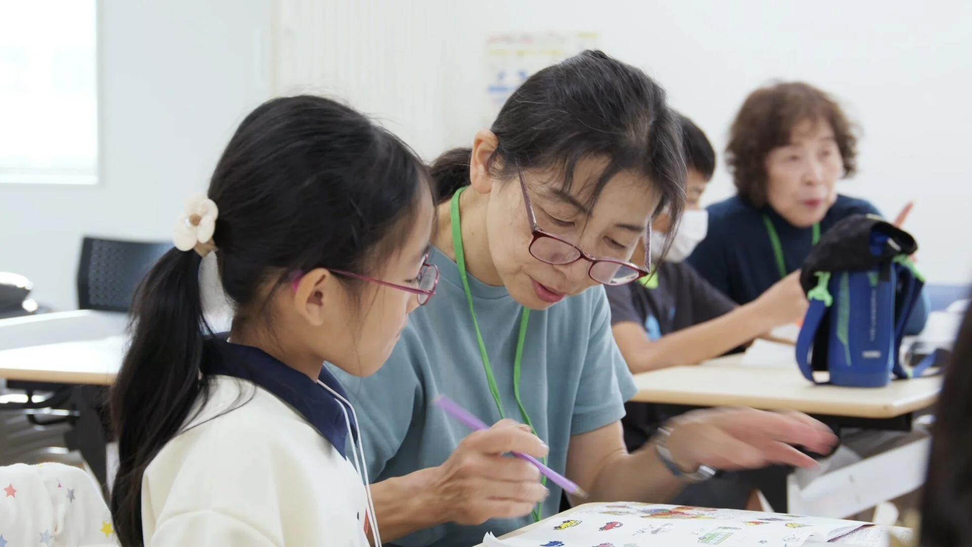 An Asian woman assisting a young Asian girl with her schoolwork in a classroom. Other students are seated at tables in the background.