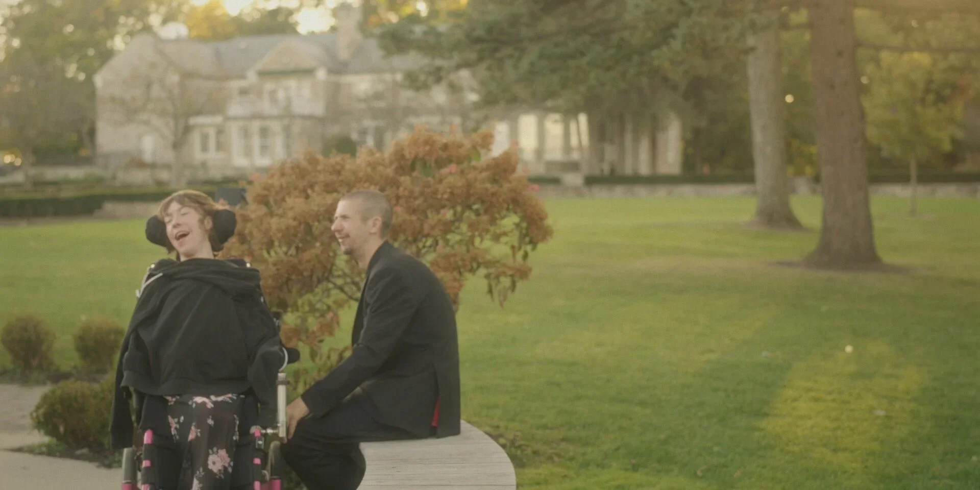 Two people sitting on a park bench, two smiling women, one in a wheelchair, the other sitting next to her, both laughing, surrounded by trees and a lawn.