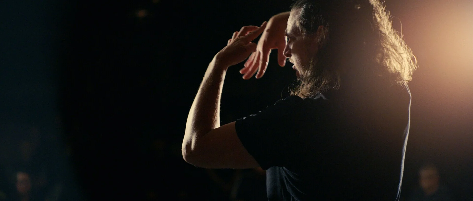 Person with long hair in black shirt, head slightly bowed, hand touching forehead, backlit with a warm glow.