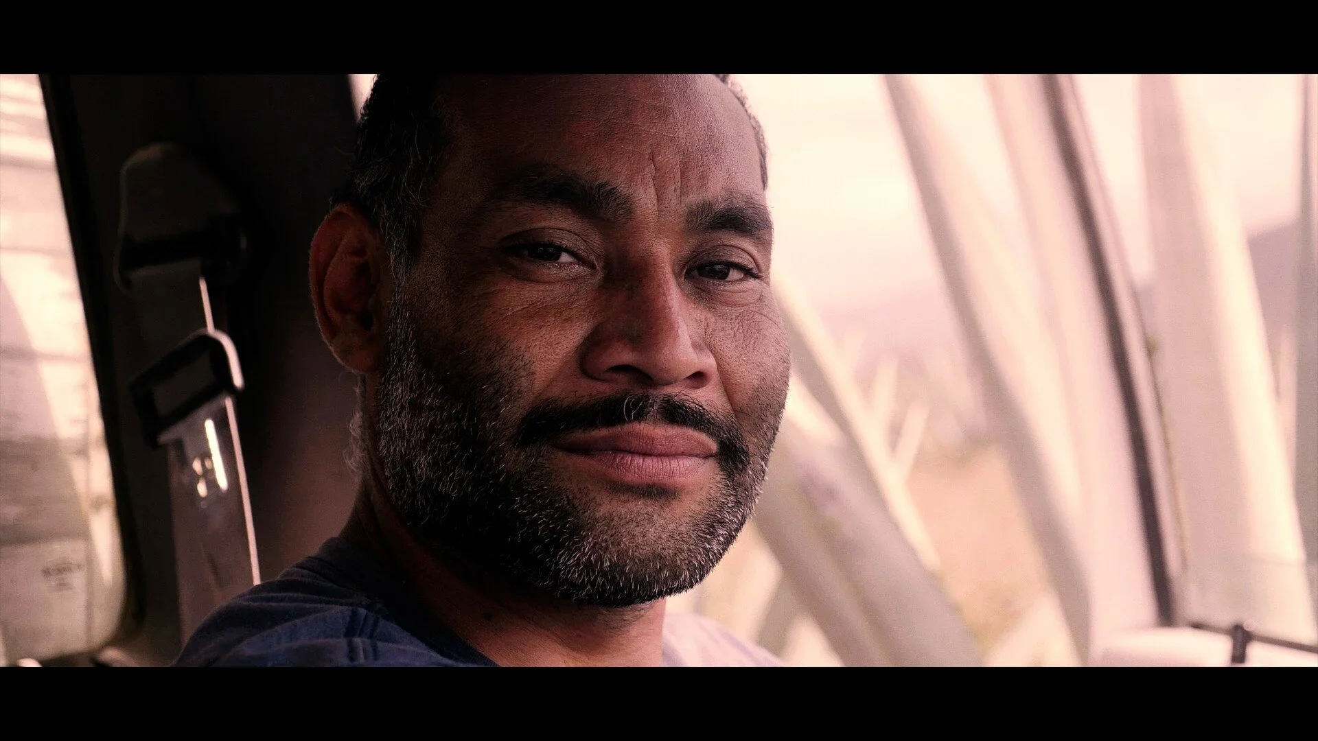 Close-up of a man with dark hair and a beard, sitting inside a vehicle with large windows, looking at the camera with a slight smile.