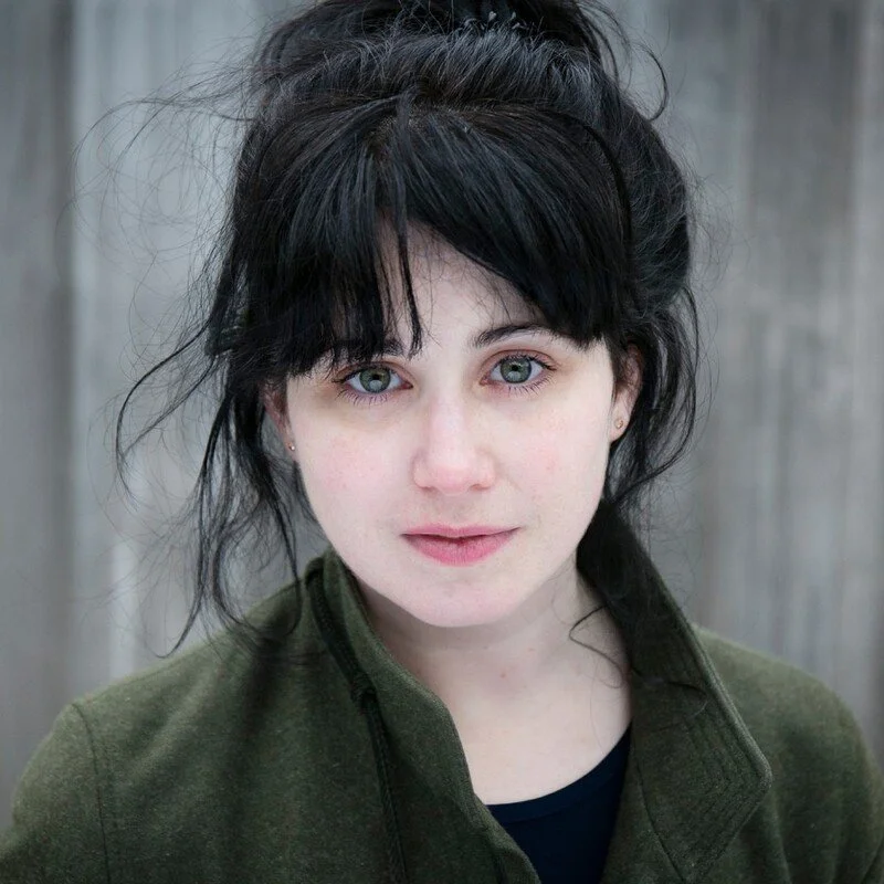 A young woman with messy black hair and light skin, looking directly at the camera with a neutral expression, standing outdoors in front of a wooden fence.