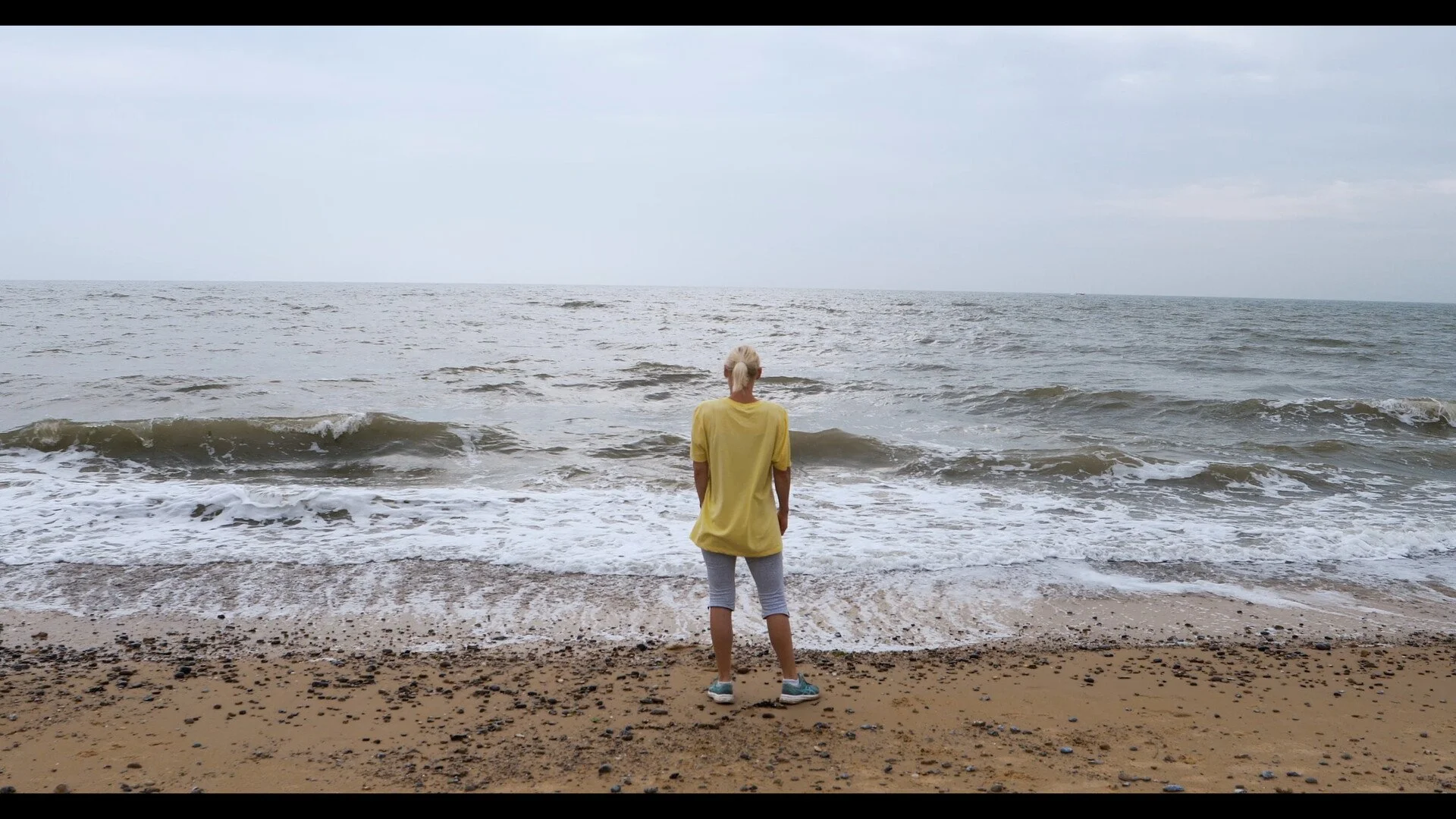 Person standing on the beach facing the ocean with waves, overcast sky