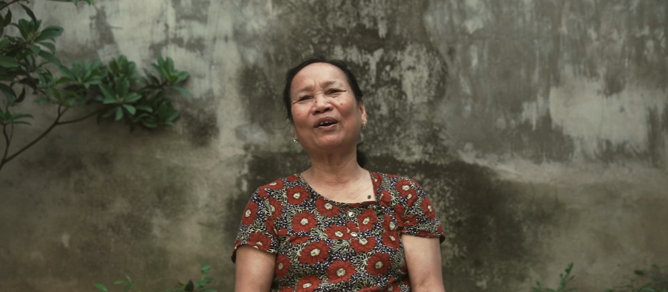 A woman standing outdoors near a weathered concrete wall with some green plants, wearing a floral patterned dress.