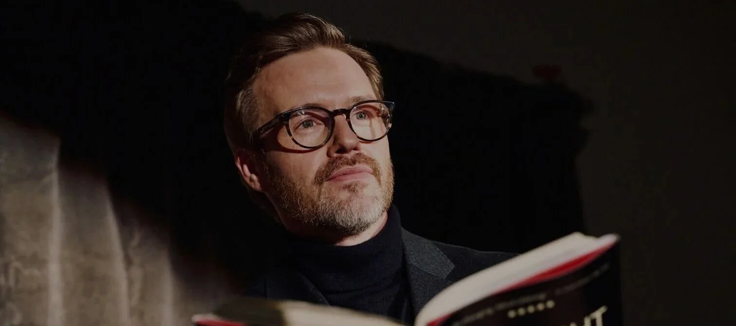A man with glasses and a beard reading a book in a dimly lit room.