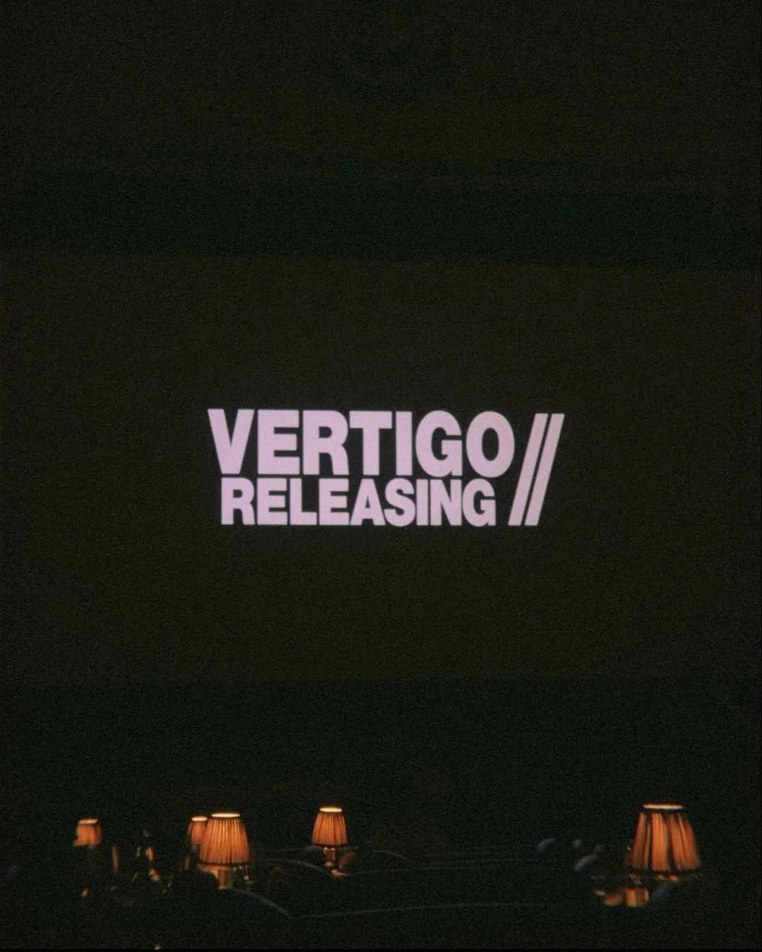 A dark theater screen shows the text 'VERTIGO RELEASING' in purple with two slashes. The bottom of the image shows several small, lit table lamps.