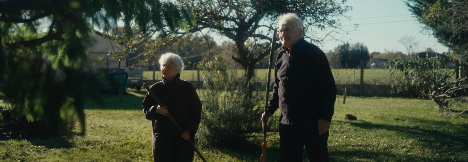 An elderly man and woman standing outdoors in a backyard, each holding gardening tools, with trees and a fence in the background.