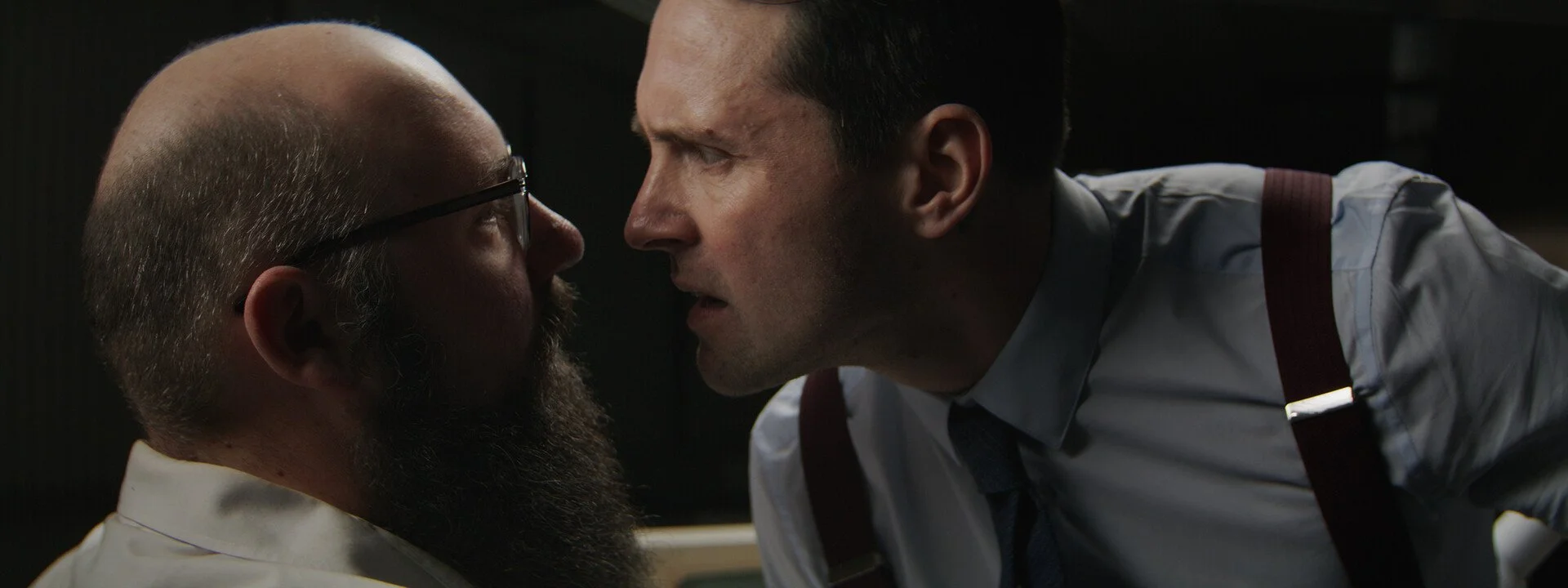 Two men with serious expressions are face to face, leaning in close, in a tense confrontation in a dimly lit setting.