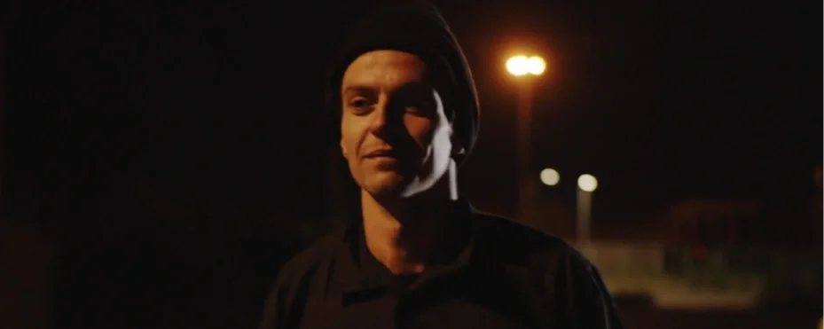 A man wearing a black beanie and dark shirt standing outdoors at night under a streetlight.