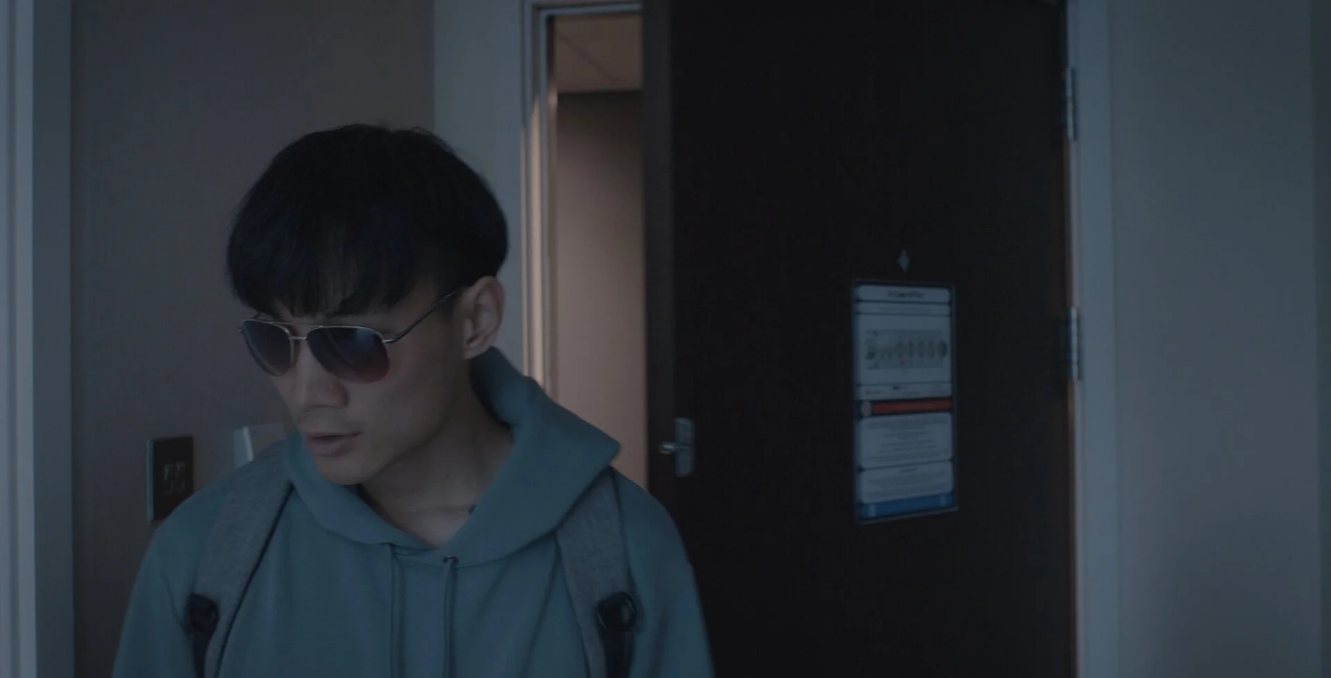 A man with black hair wearing sunglasses, a gray hoodie, and a backpack standing in front of a dark door in a hotel hallway.