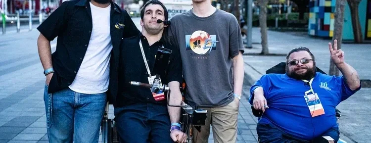 Four men posing outdoors, with one in a wheelchair making a peace sign.