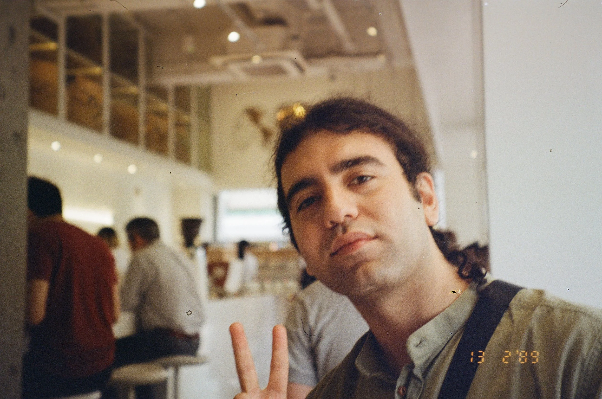 A young man with black hair and light skin making a peace sign with his fingers, smiling gently at the camera inside a cafe with blurred people and a counter in the background.