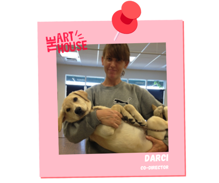 A young woman holding a playful yellow Labrador Retriever puppy inside a room with large windows. The photo is framed with a pink border, a red pushpin graphic at the top, and includes text like 'The Art House,' 'Darci,' and 'Co-Director.'