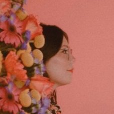 Profile of a woman with glasses and dark hair, surrounded by colorful flowers against a pink background.