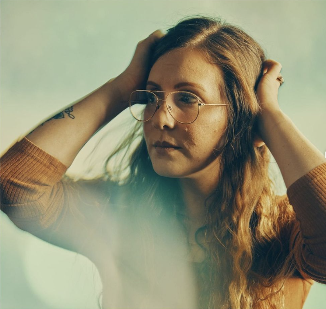 A woman with long brown hair wearing glasses and a brown top, holding her head with her hands, looking contemplative.