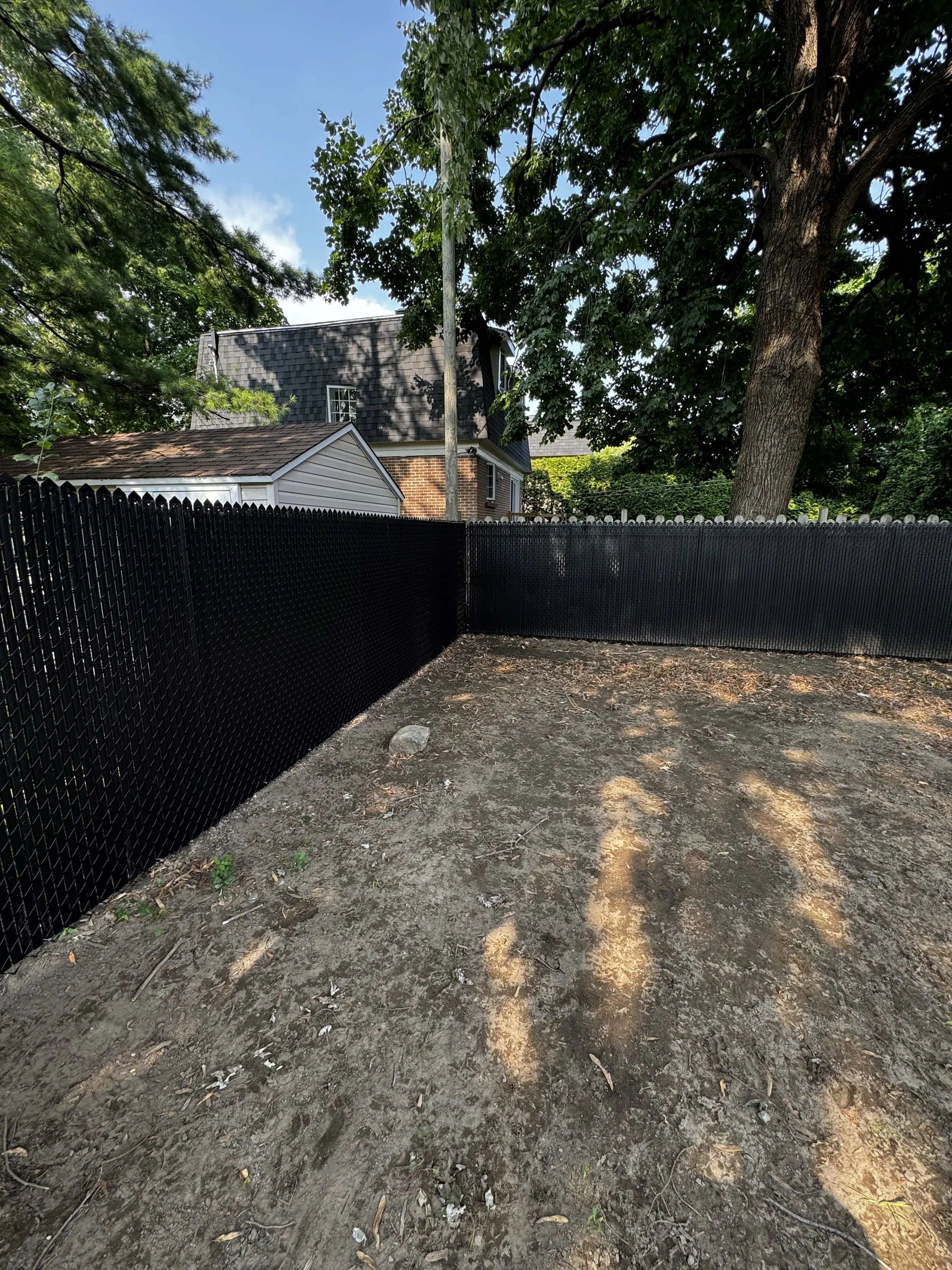 Terrain en terre avec une clôture noire en plastique ou en métal entourant un jardin ou un espace extérieur, avec des arbres et une maison en arrière-plan.