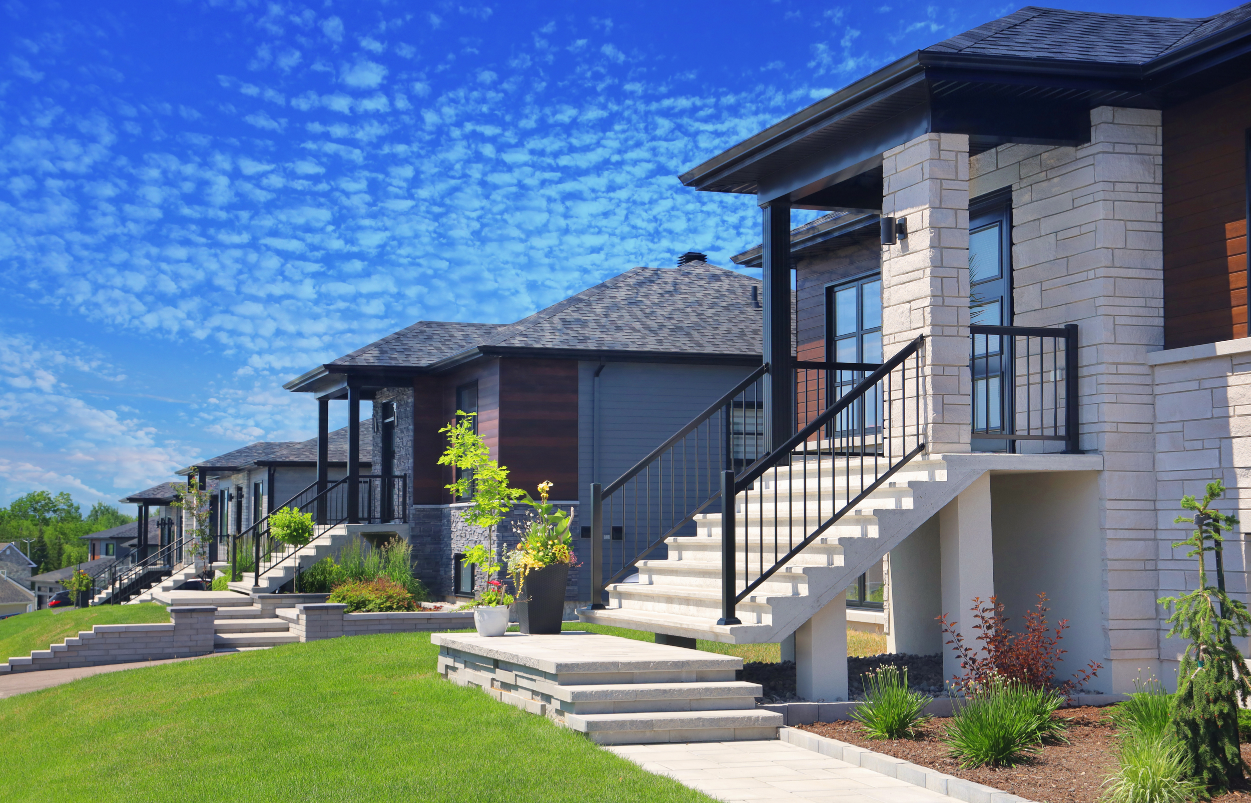 Vue d'une rangée de maisons modernes avec escaliers extérieurs, pelouse verte et ciel bleu avec des nuages.