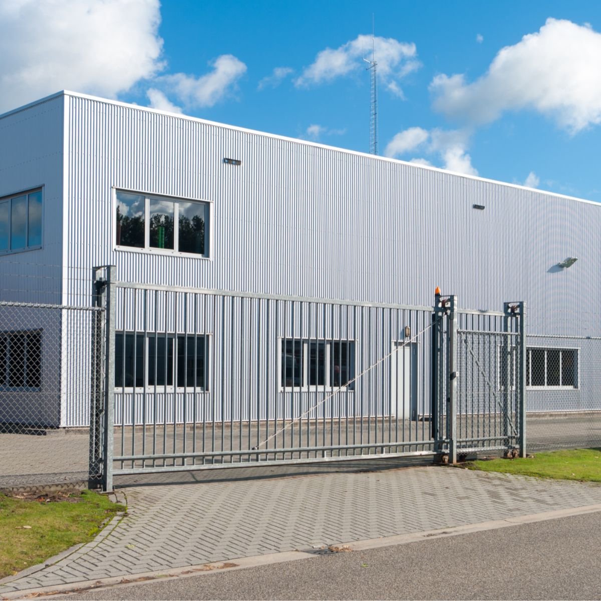 Bâtiment industriel en métal blanc avec un portail en fer et une grille, sous un ciel bleu avec quelques nuages.