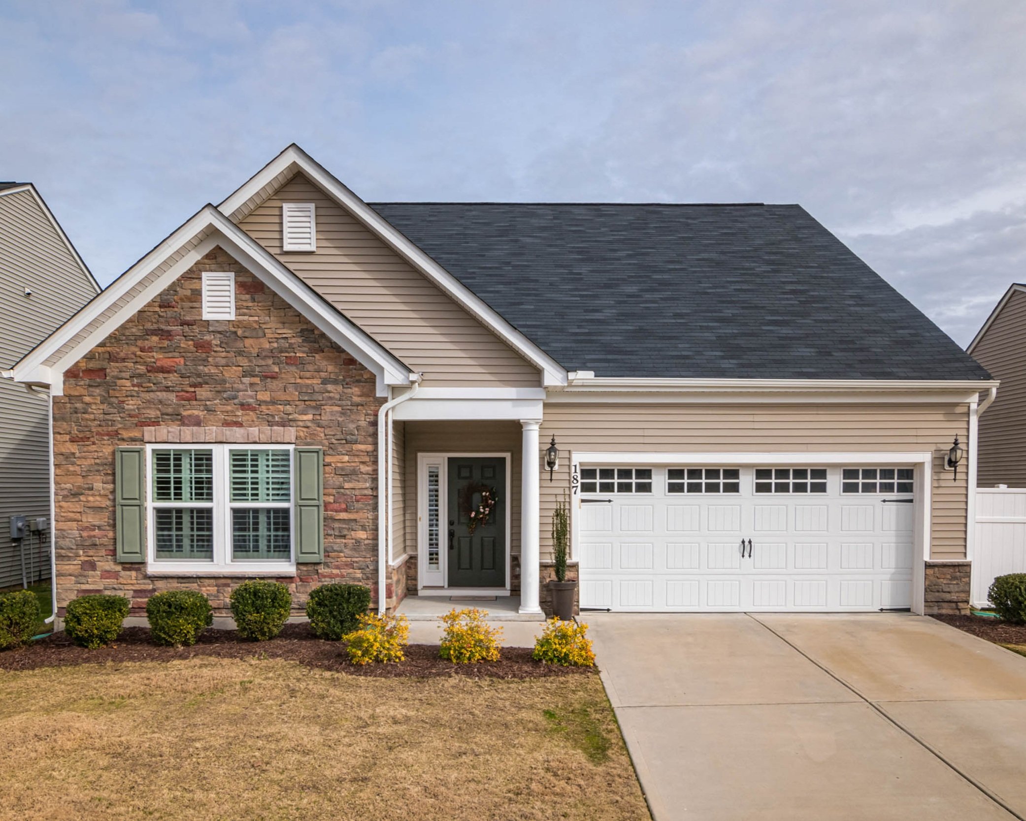 Maison moderne avec façade en pierre et en vinyl, porte d'entrée verte avec guirlande de Noël, garage double avec porte blanche, jardin avec buissons et fleurs