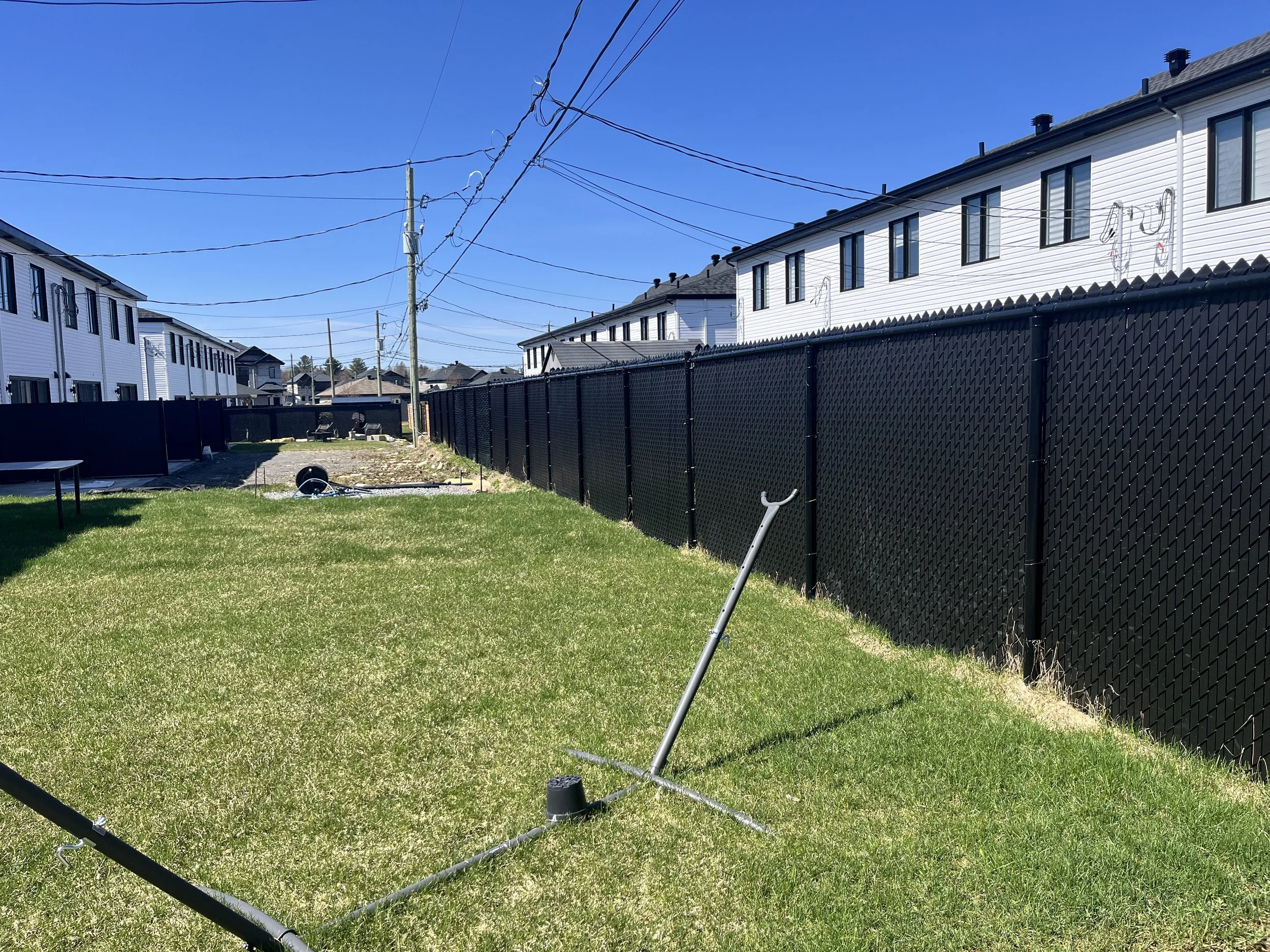 Un jardin avec une pelouse verte, une clôture noire en plastique, des maisons blanches à deux étages, un ciel bleu clair sans nuages, et des câbles électriques suspendus au-dessus.