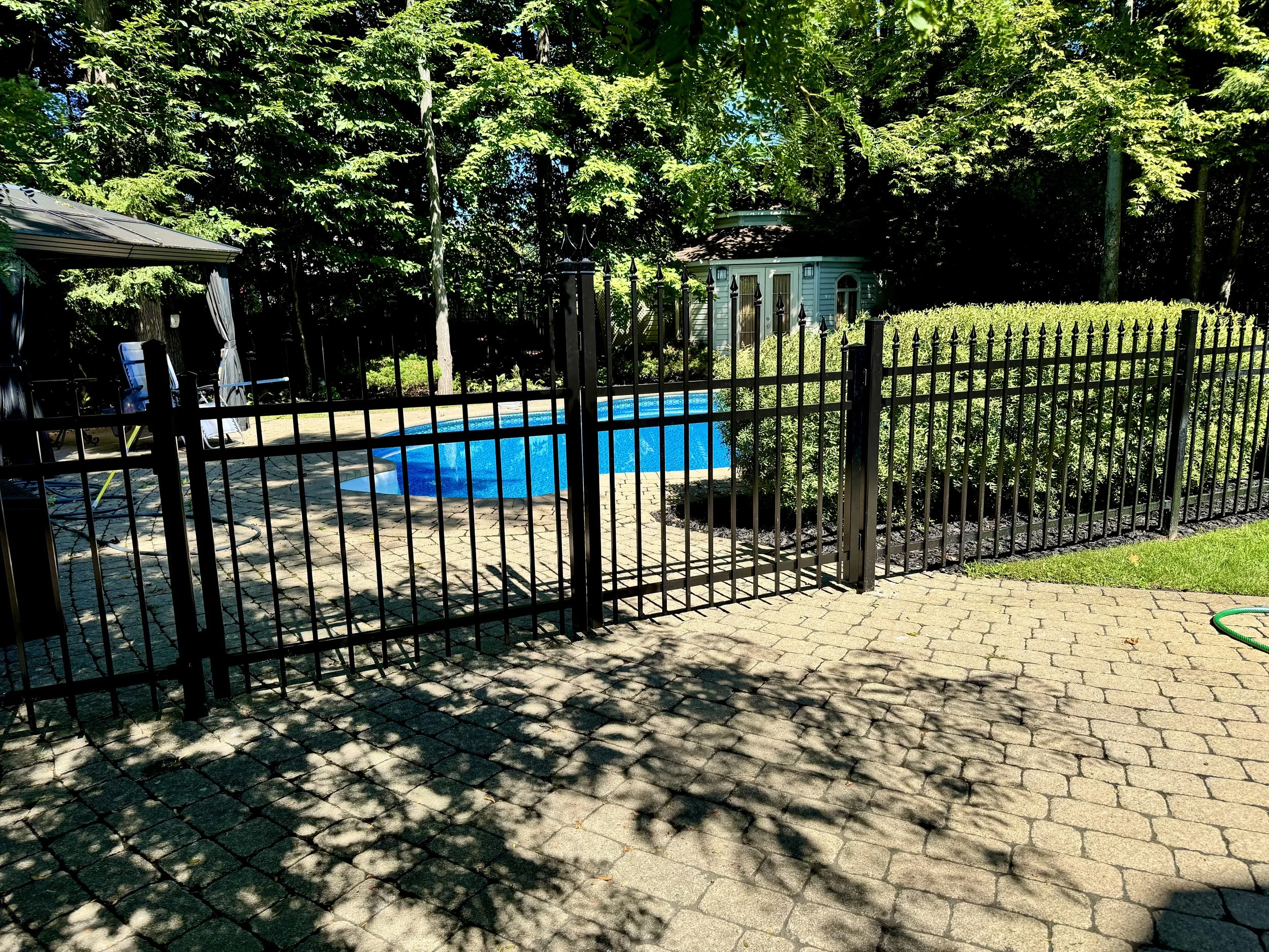 Piscine derrière une clôture noire, entourée d'arbres verts et d'une maisonette en bois bleue dans un jardin ensoleillé.