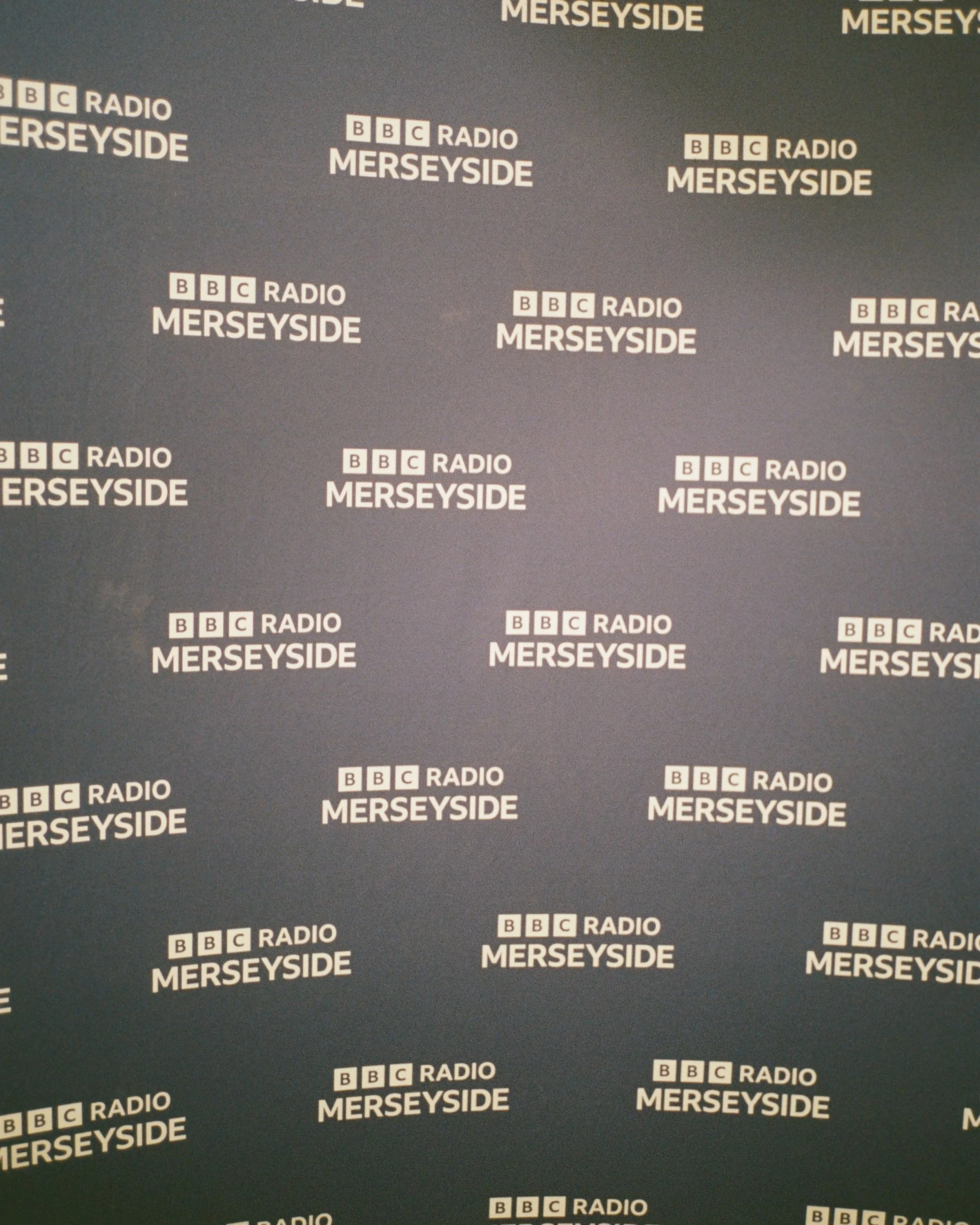 Two people standing in front of a blue backdrop with the BBC Radio Merseyside logo, posing for a photo.
