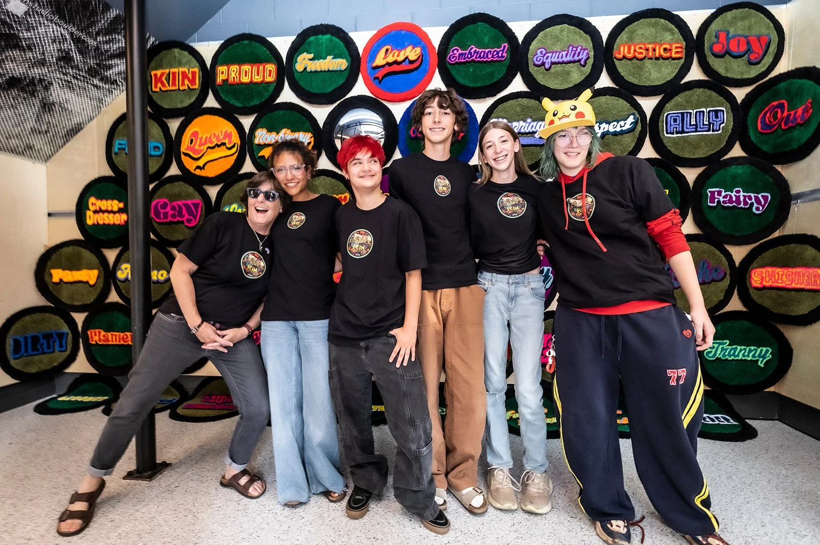 A group of five teenagers stand together smiling, with their arms around each other, in front of a wall decorated with colorful, textured art. The teens are wearing casual clothes, some with graphic T-shirts and one with a plaid shirt, and all are wearing sneakers. The art on the wall includes a large black rooster and a horse, a giant bird made of textured material, and a colorful floral-patterned horse.
