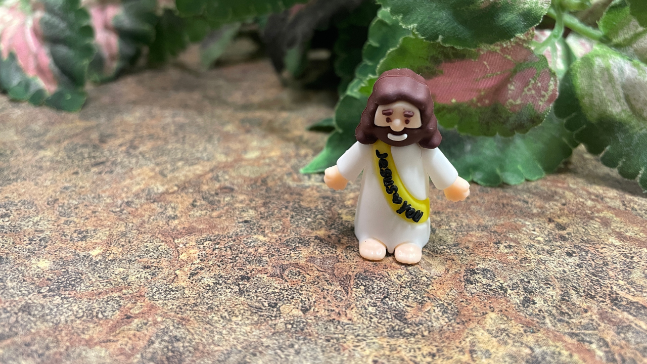 The mini Jesus Christian author Mackenzie Ryan Walters found has brown hair, a large smile, and is dressed in a white robe with a yellow sash that says, “Jesus loves you.”