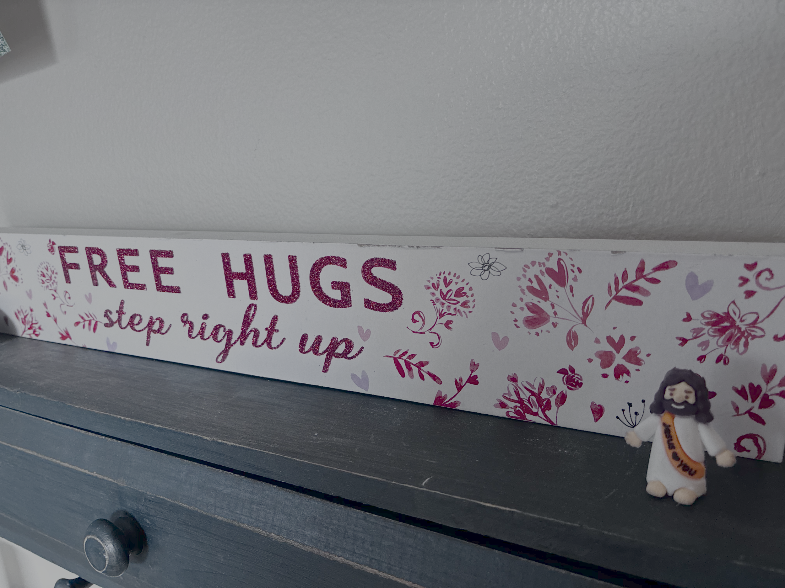 A small Jesus figurine is on the mantel next to a "free hugs" sign. It's part of a viral social media trend to "hide" toy Jesus figures as an expression of God's love in a fun way.