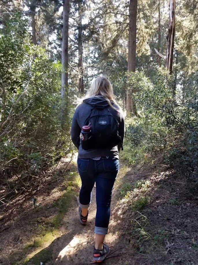 After leaving the U.S. Marines, Brittany Taga is pictured with a backpack hiking on forest trail, training for mission trip, symbolizing humility and preparation in her faith story "God gives grace to the humble"
